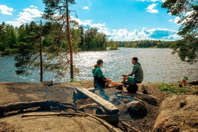 Nainen ja mies istuu nuotiopaikan penkillä kalliolla, taustalla järvi. Aurinko paistaa.